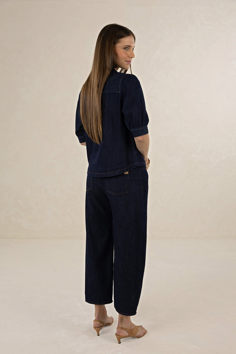 A woman with long light brown hair is shown from the back wearing a dark indigo denim two-piece set consisting of a short-sleeve top and cropped wide-leg pants, paired with beige high-heeled sandals, against a neutral cream studio backdrop.