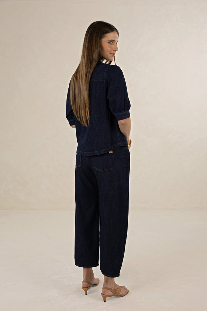 A woman with long light brown hair is shown from the back wearing a dark indigo denim two-piece set consisting of a short-sleeve top and cropped wide-leg pants, paired with beige high-heeled sandals, against a neutral cream studio backdrop.