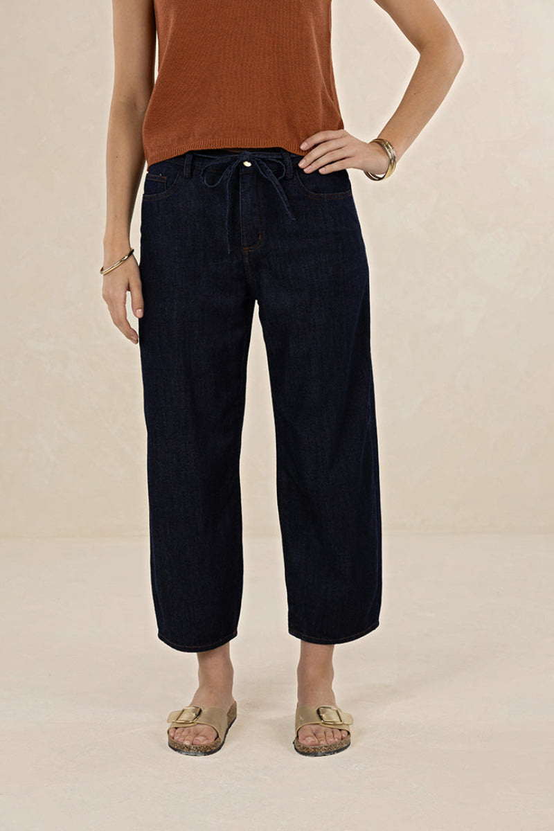 A model wearing dark indigo cropped drawstring jeans, paired with a rust-colored ribbed tank top and beige slide sandals, standing against a textured beige studio backdrop.