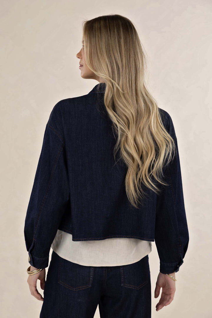 A woman shown from the back wearing a dark navy cropped denim jacket over a cream top and dark jeans, set against a plain beige studio background.