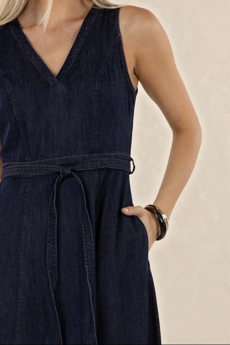 A close-up of a woman wearing a dark navy blue denim-like sleeveless dress with a V-neckline, self-tie waist belt, and side pockets, set against a plain beige background.