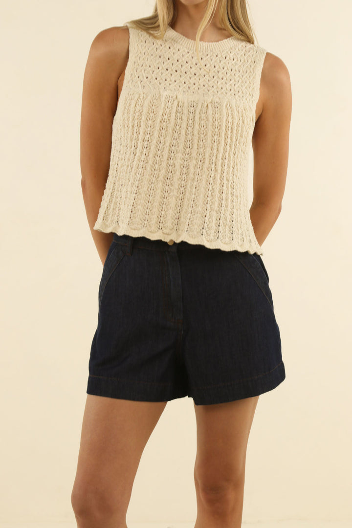 A woman wearing a sleeveless cream-colored knit top with a scalloped hem and dark navy blue denim shorts, standing against a solid light beige background.