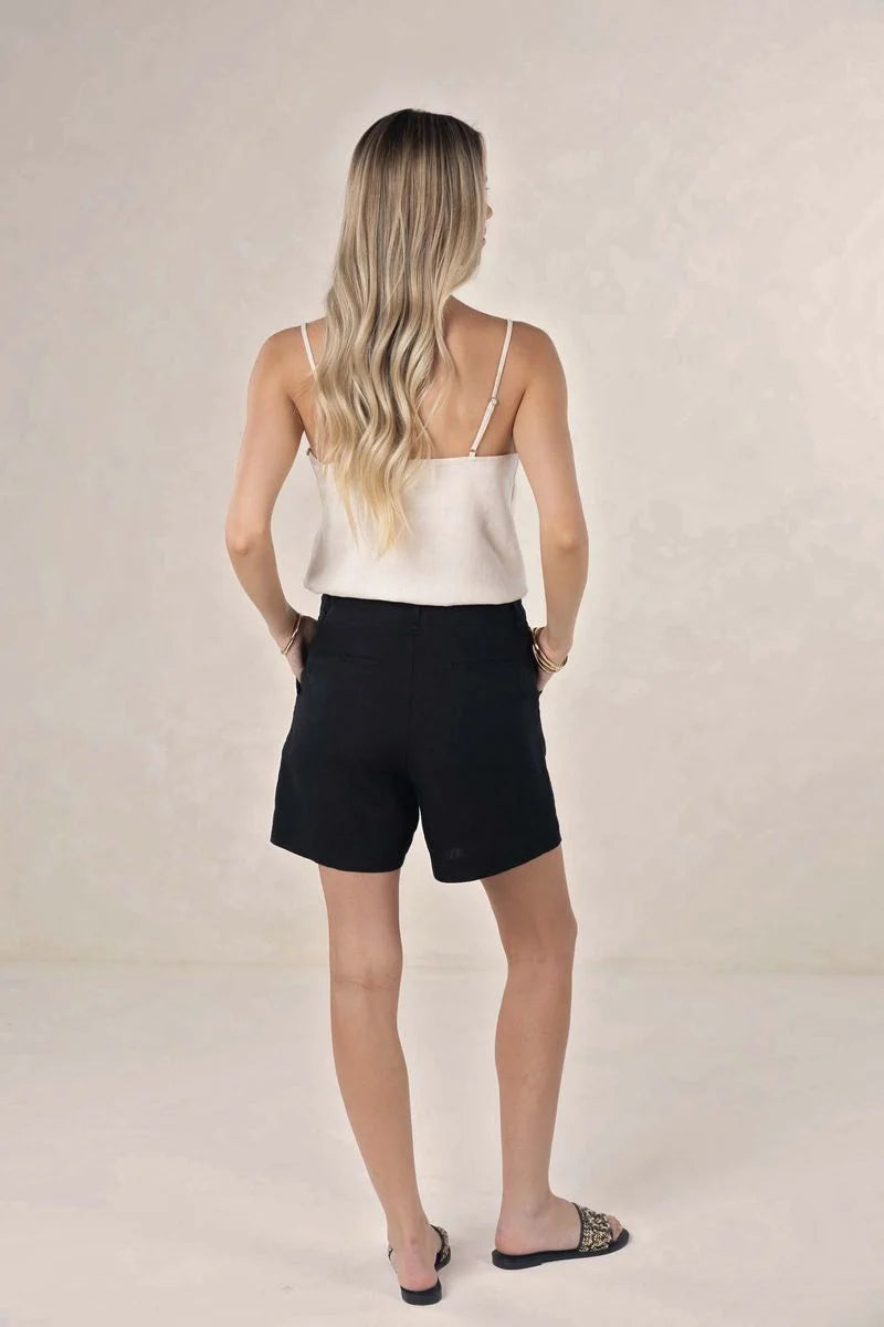 A woman with blonde hair is shown from behind wearing black shorts and a white camisole top, standing against a neutral beige studio background.