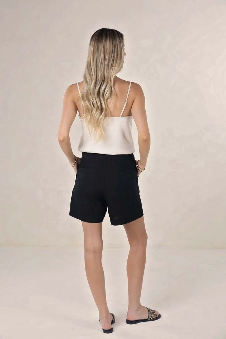 A woman with blonde hair is shown from behind wearing black shorts and a white camisole top, standing against a neutral beige studio background.