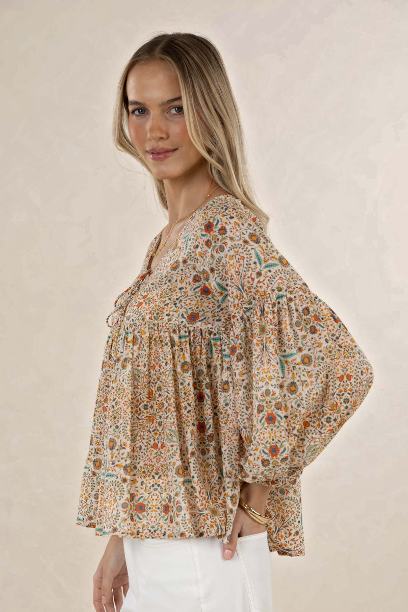 A woman modeling a bohemian-style long-sleeve blouse with a dense multicolor floral and geometric print on a beige background, paired with white pants, set against a neutral beige textured wall.