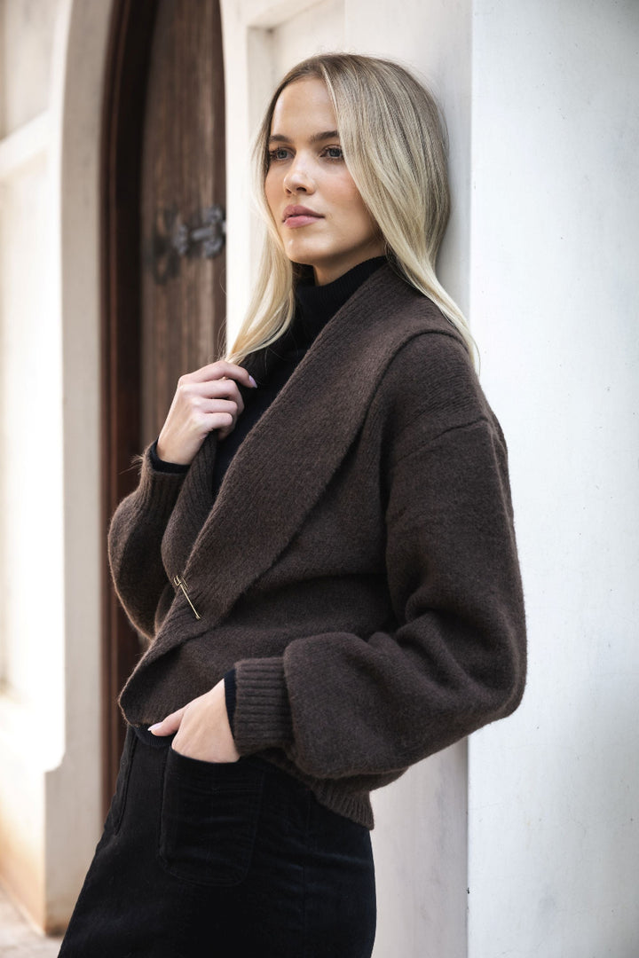 Woman wearing a brown coat standing against a white wall.
