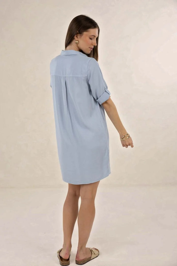 A woman wearing a light blue oversized button-up shirt dress, shown from a side-back angle against a neutral beige studio background.