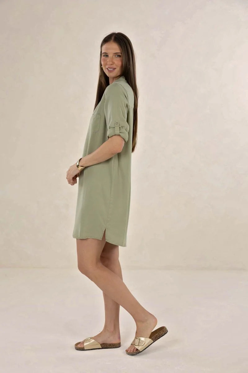 A woman modeling a sage green short-sleeve tunic dress with rolled cuffs, paired with metallic gold slide sandals, set against a plain beige studio backdrop.