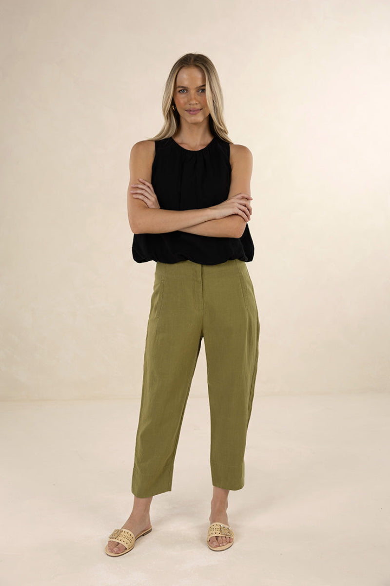 A woman modeling a sleeveless black top and olive green cropped pants with beige slide sandals, standing against a plain beige studio background.