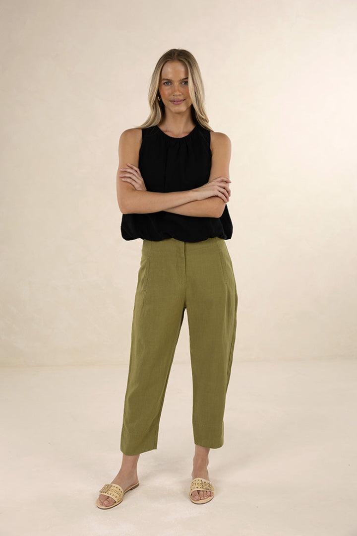 A woman modeling a sleeveless black top and olive green cropped pants with beige slide sandals, standing against a plain beige studio background.