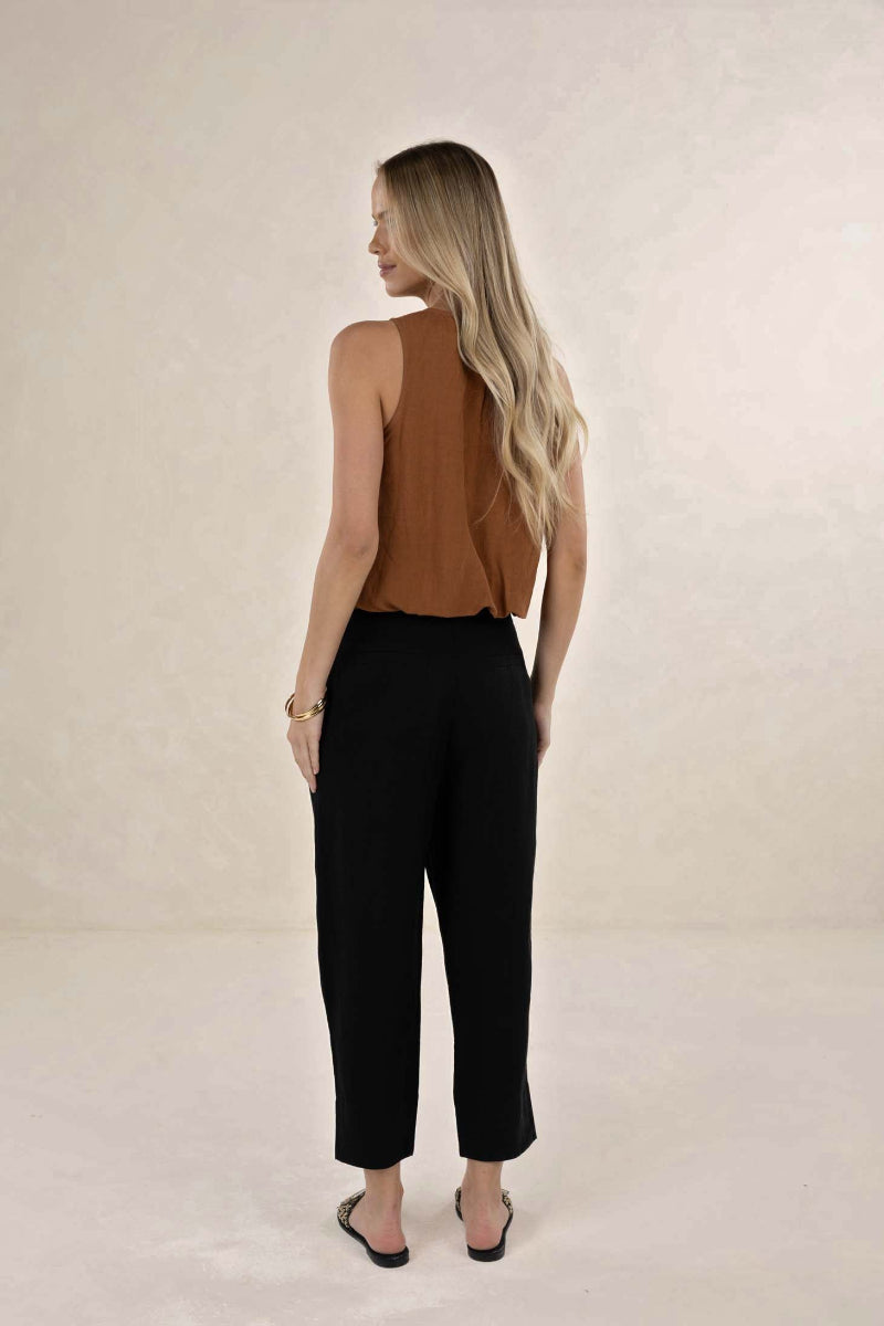 A woman is shown from behind wearing a sleeveless brown top and black cropped pants, standing against a neutral beige studio backdrop.