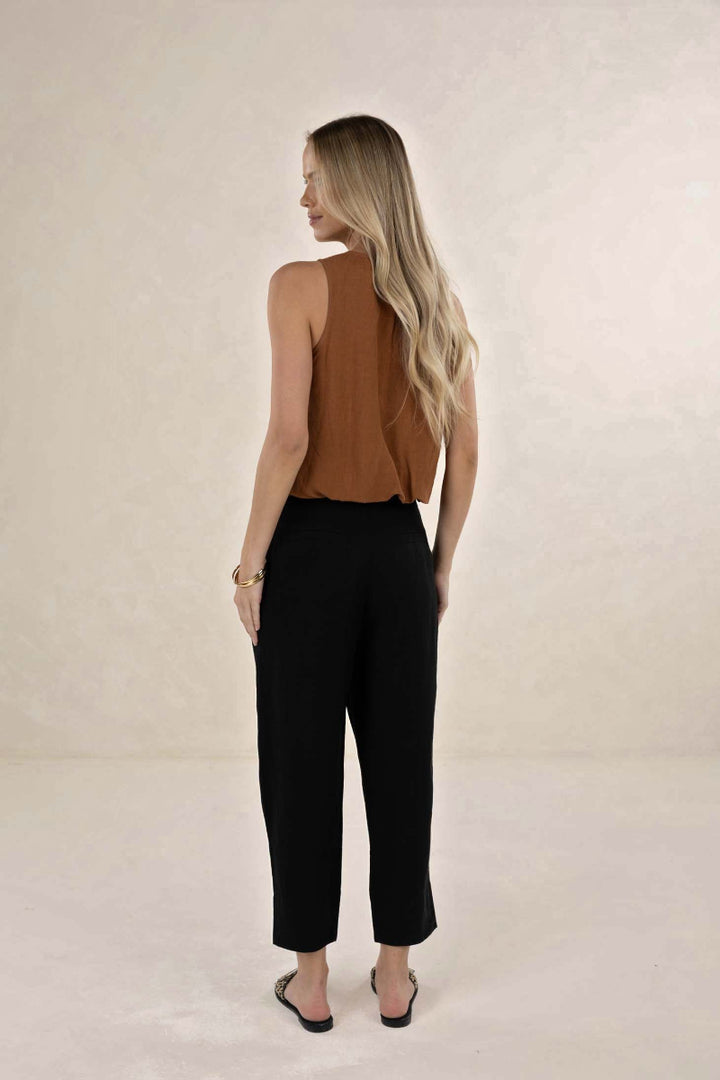 A woman is shown from behind wearing a sleeveless brown top and black cropped pants, standing against a neutral beige studio backdrop.