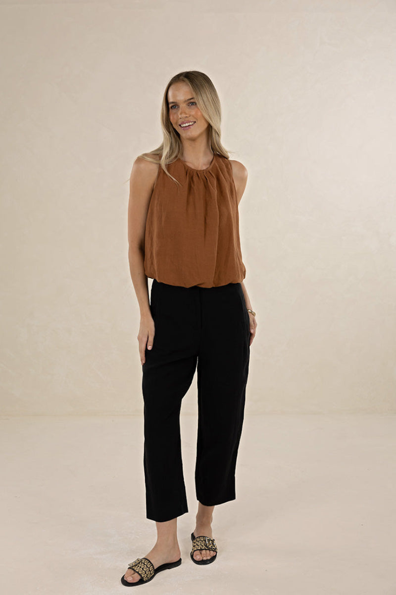 A woman modeling a sleeveless rust-brown top and black cropped pants with patterned slide sandals, standing against a plain beige studio backdrop.