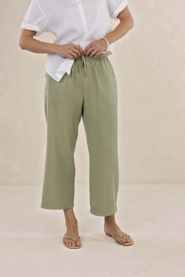 A person wearing light sage green cropped drawstring pants with side pockets, styled with a white short-sleeve button-up shirt and beige flat sandals against a warm beige studio background.