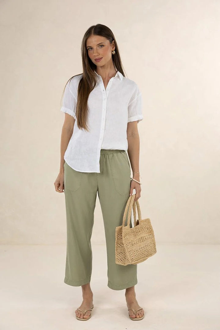 Woman wearing a white shirt and green pants, holding a woven bag on a beige background