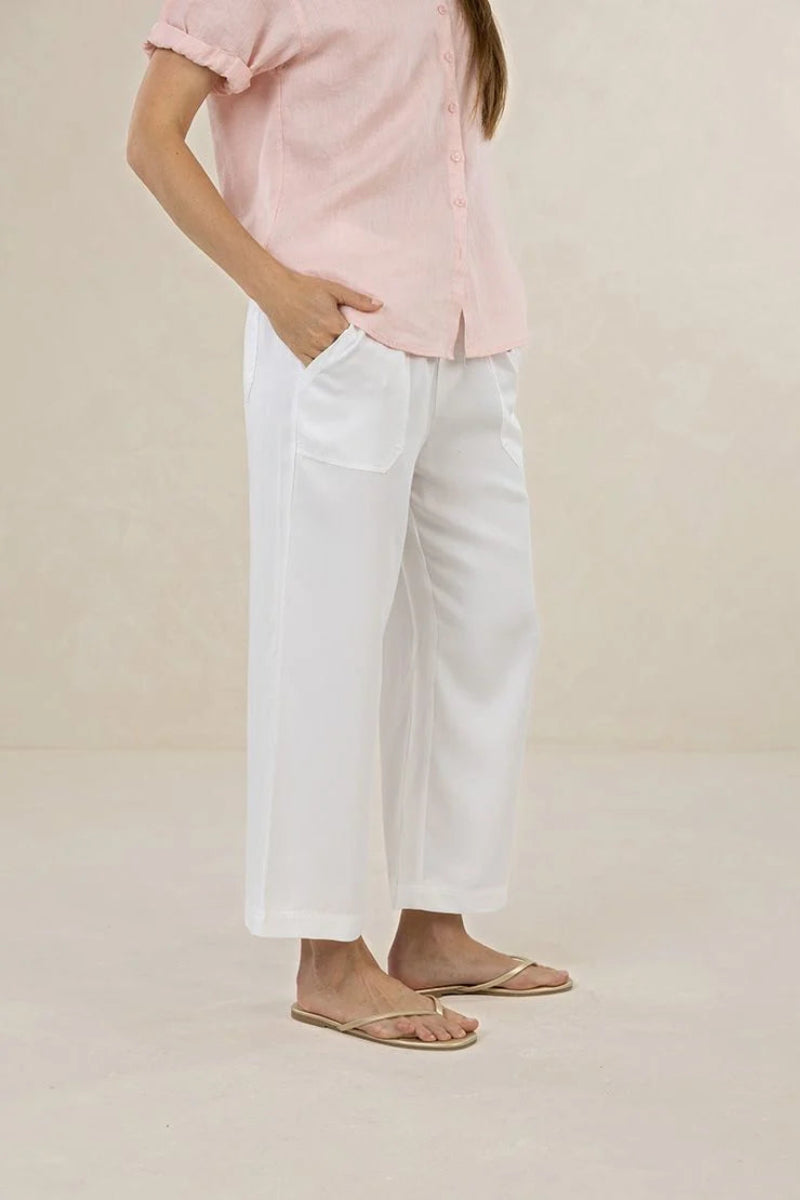 A model wearing white wide-leg cropped pants with side pockets, paired with a light pink short-sleeve button-up shirt and beige flat sandals, standing against a neutral beige background.