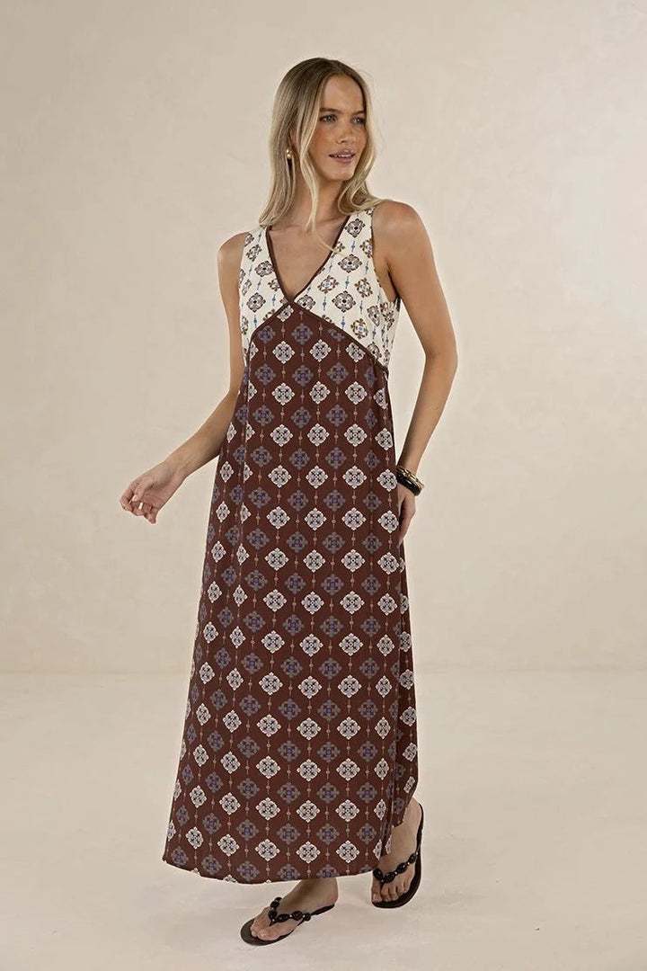 A woman modeling a sleeveless maxi dress with a brown and cream geometric pattern, featuring a white lace-up bodice, against a plain beige studio background.