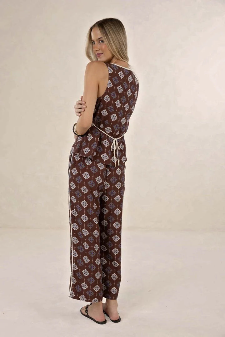 A woman modeling a sleeveless brown jumpsuit with a beige geometric pattern and a matching tie belt, shown from the back against a neutral studio backdrop.