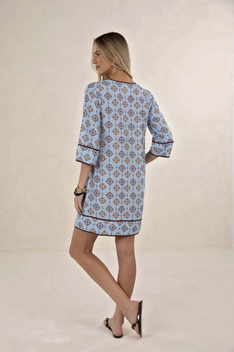 A woman modeling a light blue tunic dress with a brown and beige geometric pattern, shown from the back against a neutral beige studio backdrop.