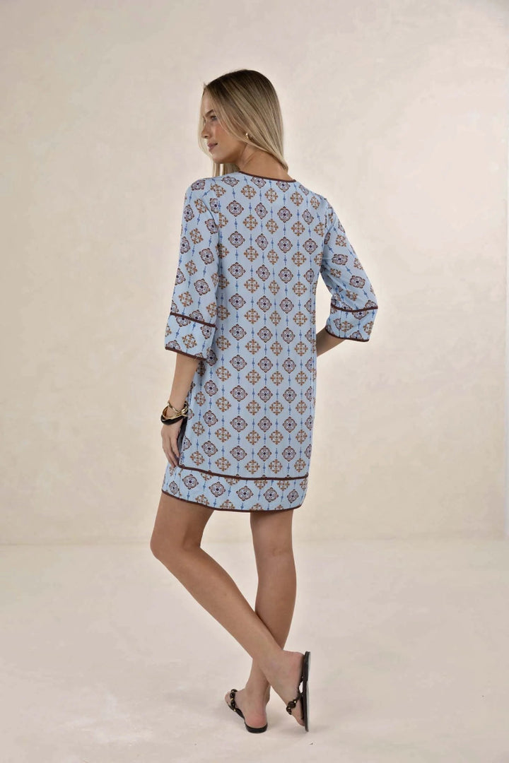 A woman modeling a light blue tunic dress with a brown and beige geometric pattern, shown from the back against a neutral beige studio backdrop.