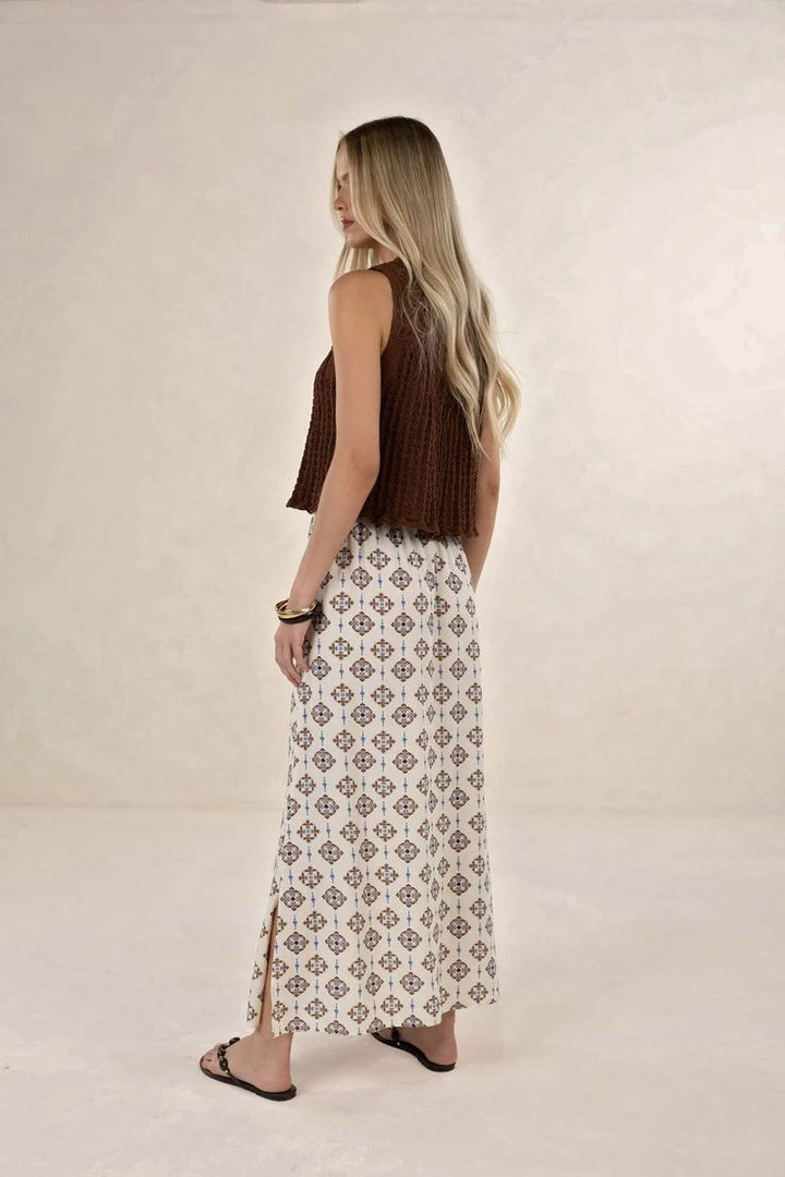 A woman is shown from the back wearing a sleeveless brown knit top and a long white maxi skirt with a brown geometric pattern, standing against a neutral beige studio background.