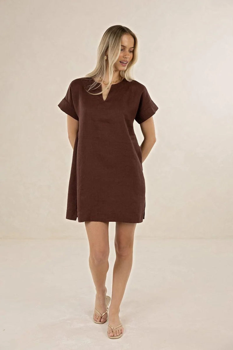 A woman modeling a short-sleeve brown mini dress with a split V-neckline, standing against a plain beige studio backdrop.