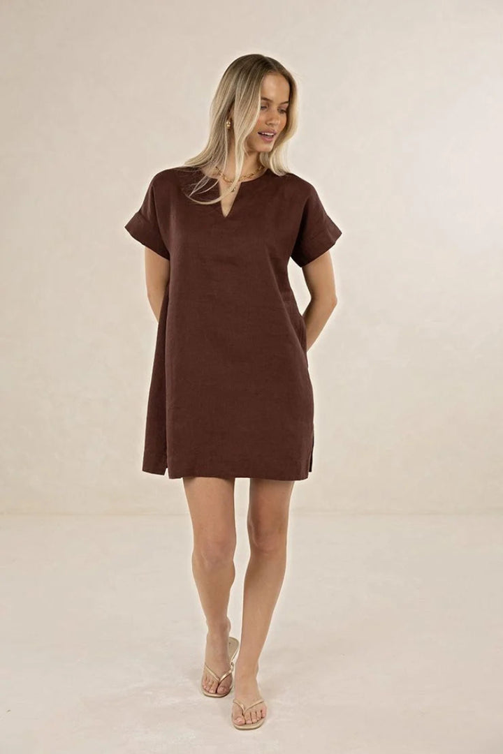 A woman modeling a short-sleeve brown mini dress with a split V-neckline, standing against a plain beige studio backdrop.
