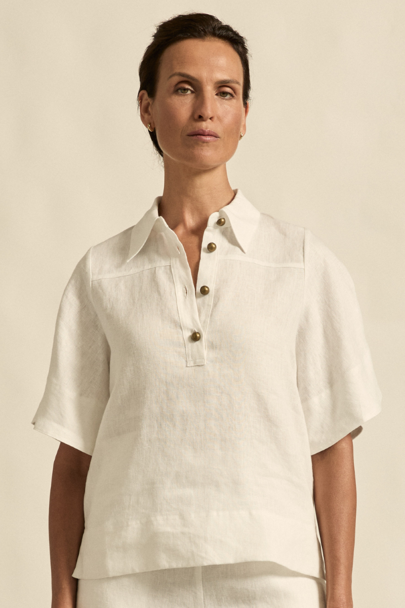 This front close-up image of the Insinuate Top in Porcelain highlights the crisp collar, delicate flutter sleeves, and brass domed buttons with refined tailoring details.
