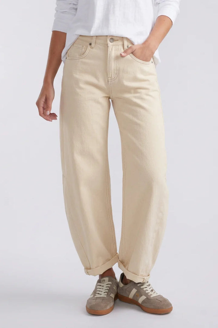 A person wearing loose, light beige wide-leg jeans with a white top and taupe sneakers, shown from the waist down against a light gray studio background.
