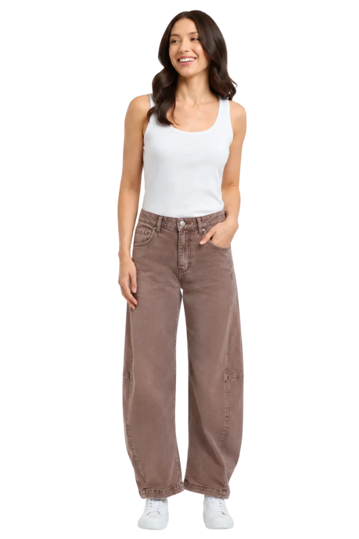 A woman modeling a pair of wide-leg brown jeans with a white ribbed tank top and white sneakers, standing against a plain white background.