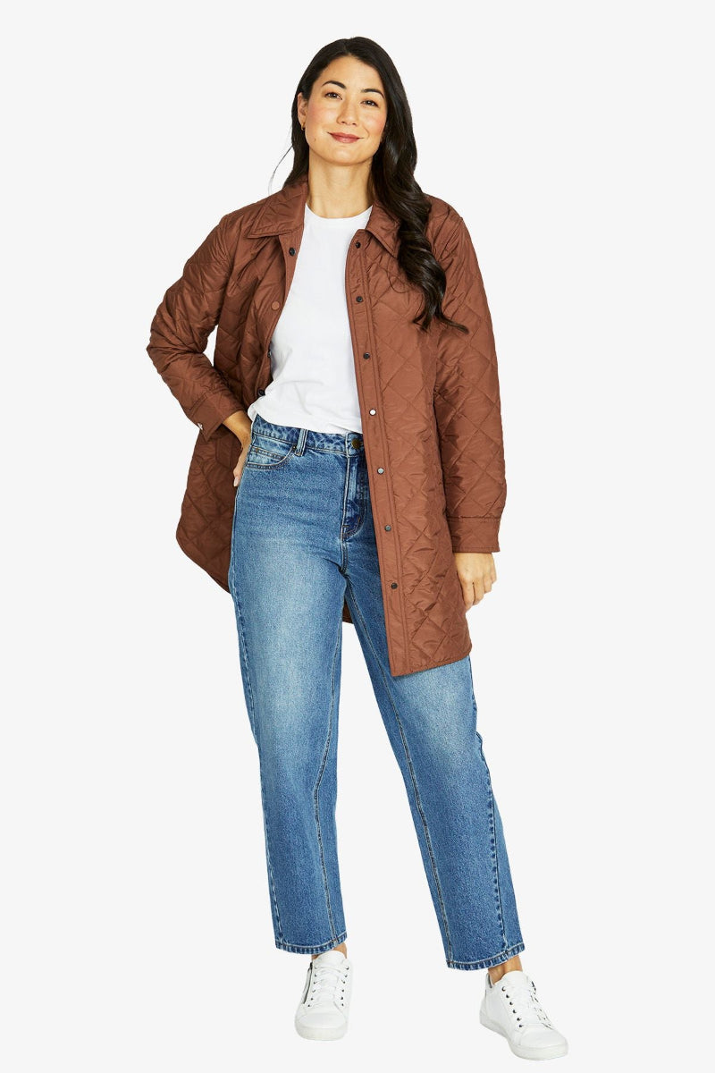 Woman wearing a brown quilted jacket, white shirt, blue jeans, and white sneakers on a white background