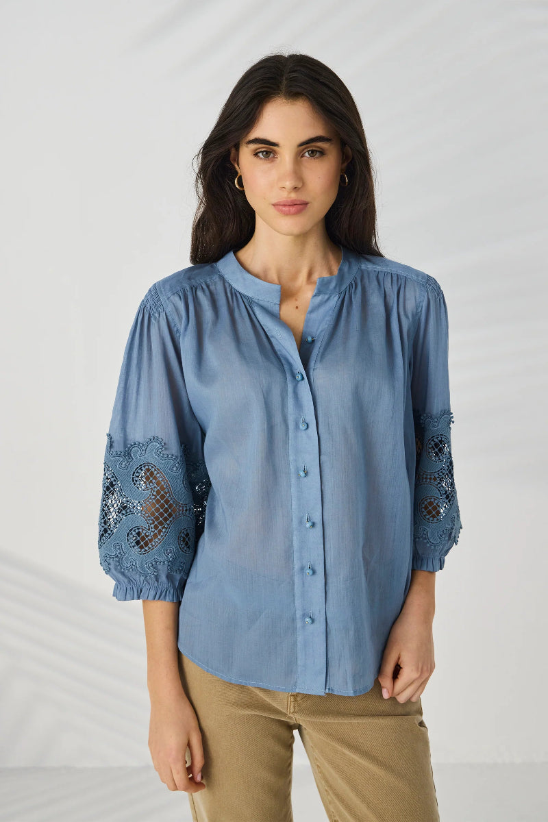 A woman modeling a light blue button-up blouse with three-quarter sleeves and lace detailing on the cuffs, paired with khaki pants, against a light gray background.