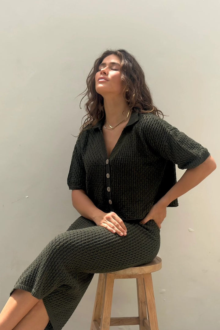 Woman wearing a dark green textured outfit sitting on a wooden stool against a plain background