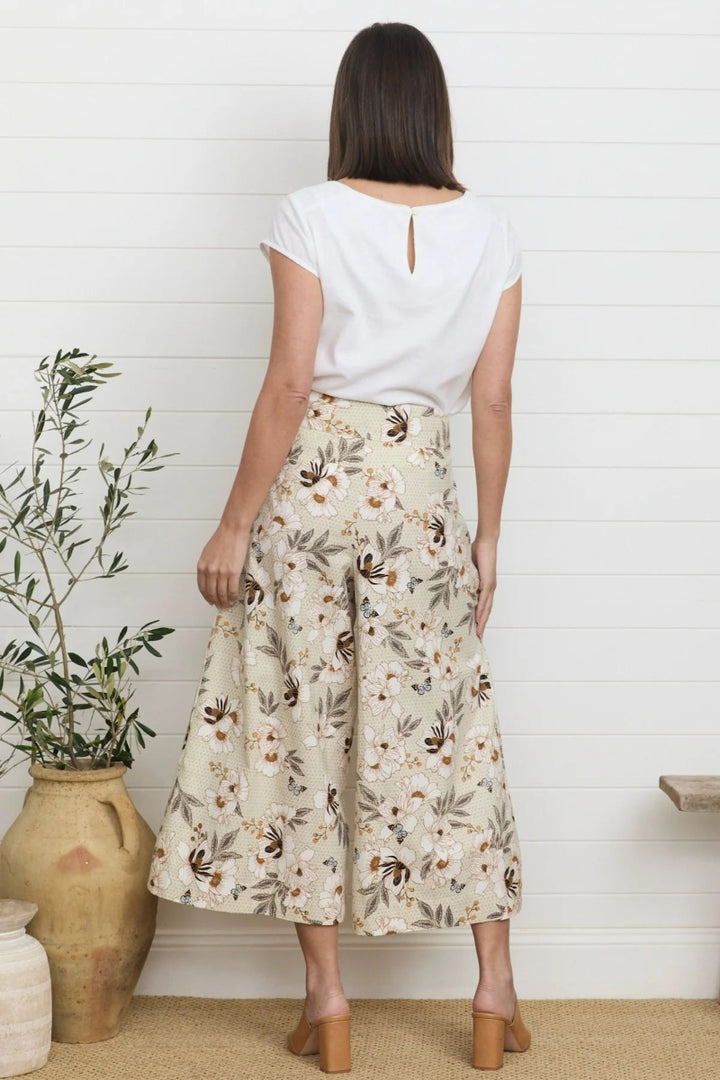 Woman wearing a white top and floral skirt standing in a room with a plant and wooden floor.