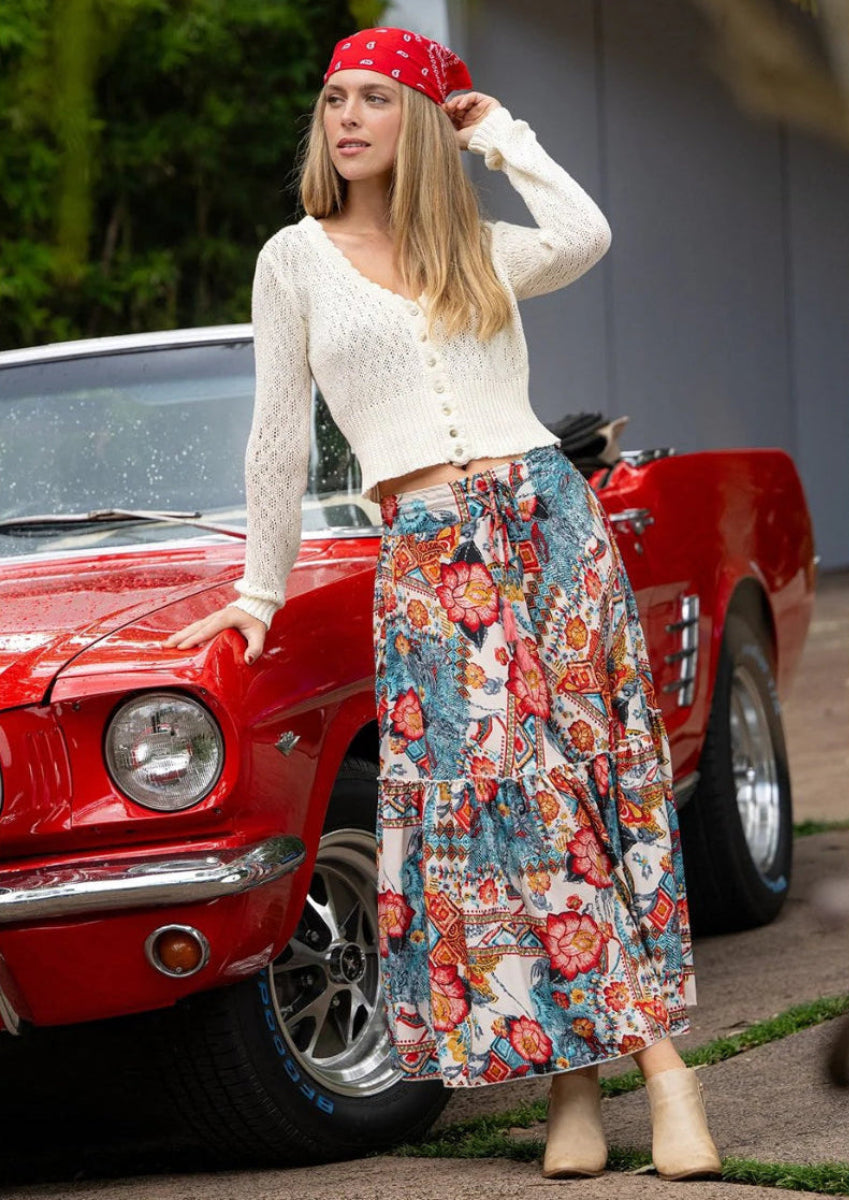 Woman in cream cardigan and frill maxi skirt standing beside a red car.