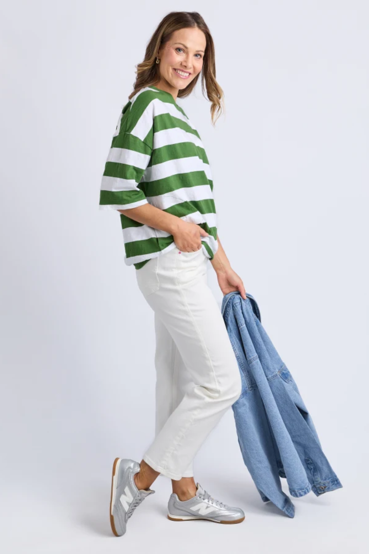 Woman wearing a green and white striped sweater with white pants, holding a denim jacket on a light gray background