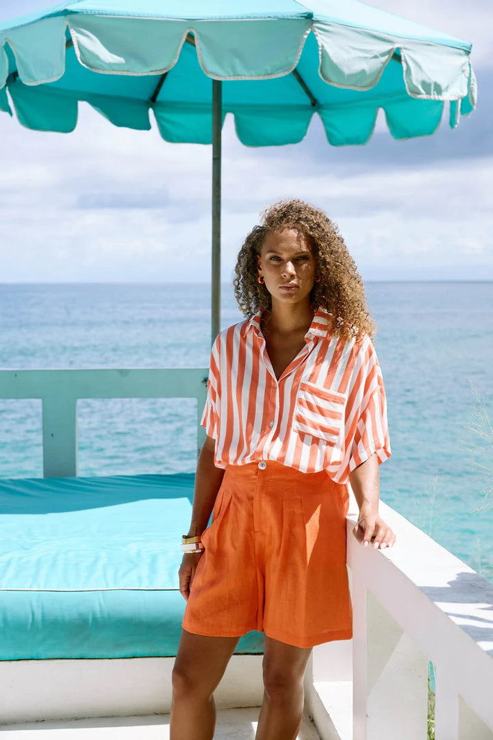 Woman in striped shirt and orange shorts standing under a teal umbrella by the ocean.