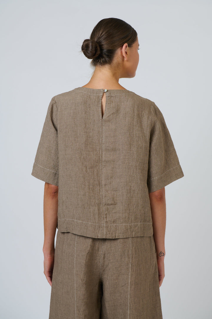 A woman shown from the back wearing a taupe linen short-sleeve top and matching trousers against a light gray studio background.
