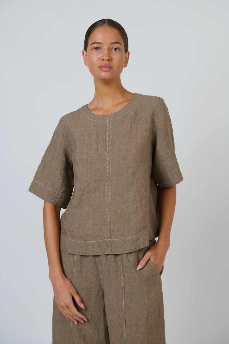 A woman modeling a matching taupe linen two-piece set consisting of a short-sleeve top with a round neckline and pleated front detail, paired with matching wide-leg trousers, against a light gray studio background.