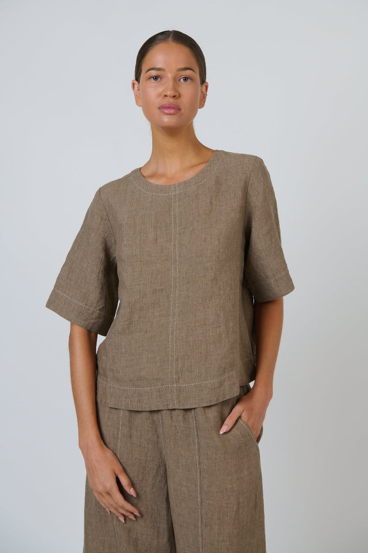 A woman modeling a matching taupe linen two-piece set consisting of a short-sleeve top with a round neckline and pleated front detail, paired with matching wide-leg trousers, against a light gray studio background.