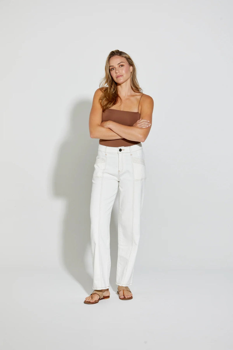 A woman stands against a light gray studio background wearing white straight-leg jeans, a brown strapless tube top, and brown sandals, with her arms crossed.