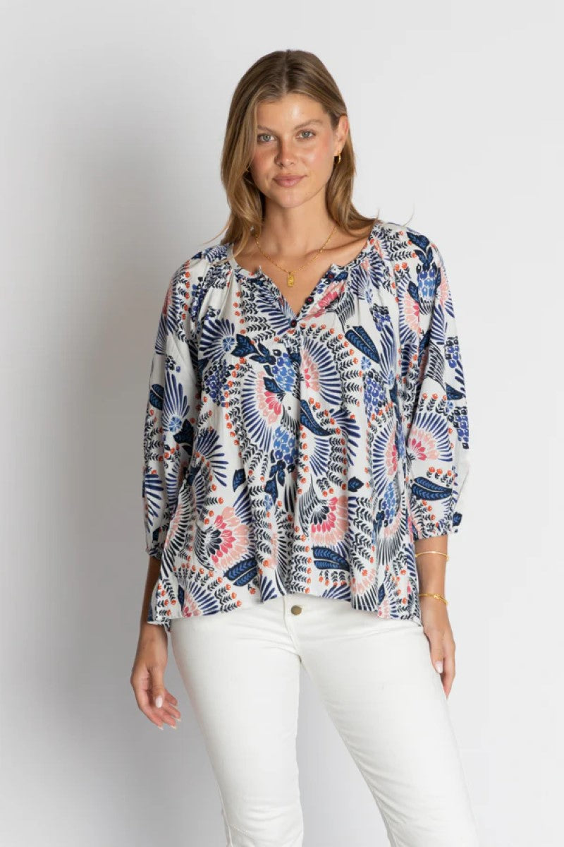 Woman wearing a patterned blouse and white pants on a plain background