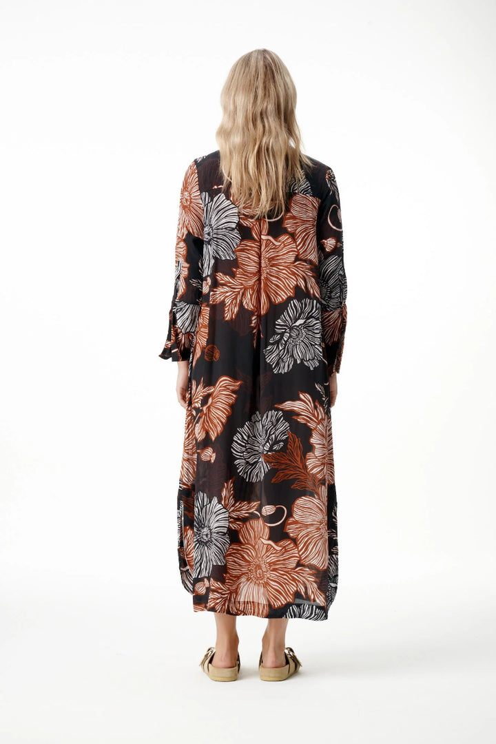 A woman viewed from behind wearing a long-sleeved maxi dress with a black base and a large floral pattern in terracotta, rust, and gray, paired with tan flat sandals, against a plain white studio background.