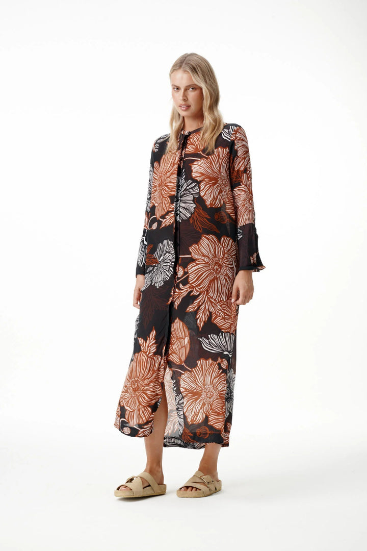 A woman modeling a long-sleeved maxi dress with a black base and a large floral print in terracotta orange, brown, and gray, paired with beige slide sandals against a white studio background.
