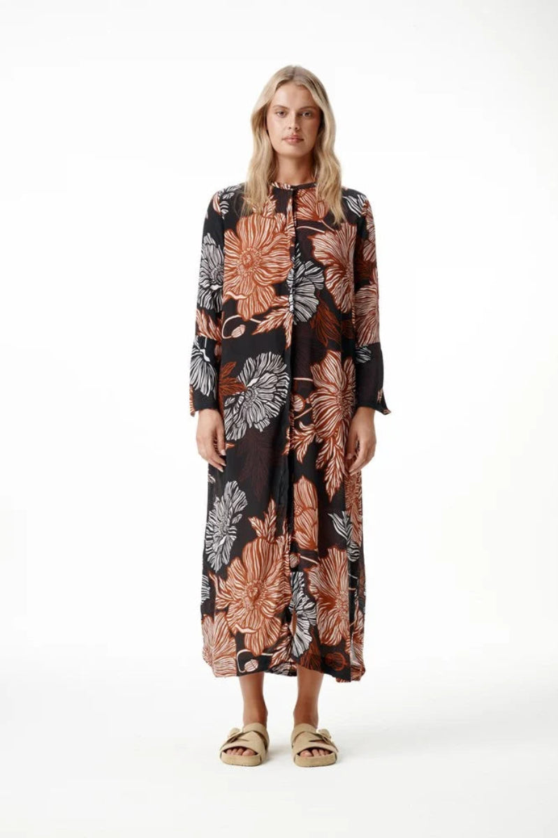 A woman modeling a long-sleeved midi dress with a black base and a large floral pattern in terracotta, rust, and gray, paired with beige slide sandals against a white studio background.
