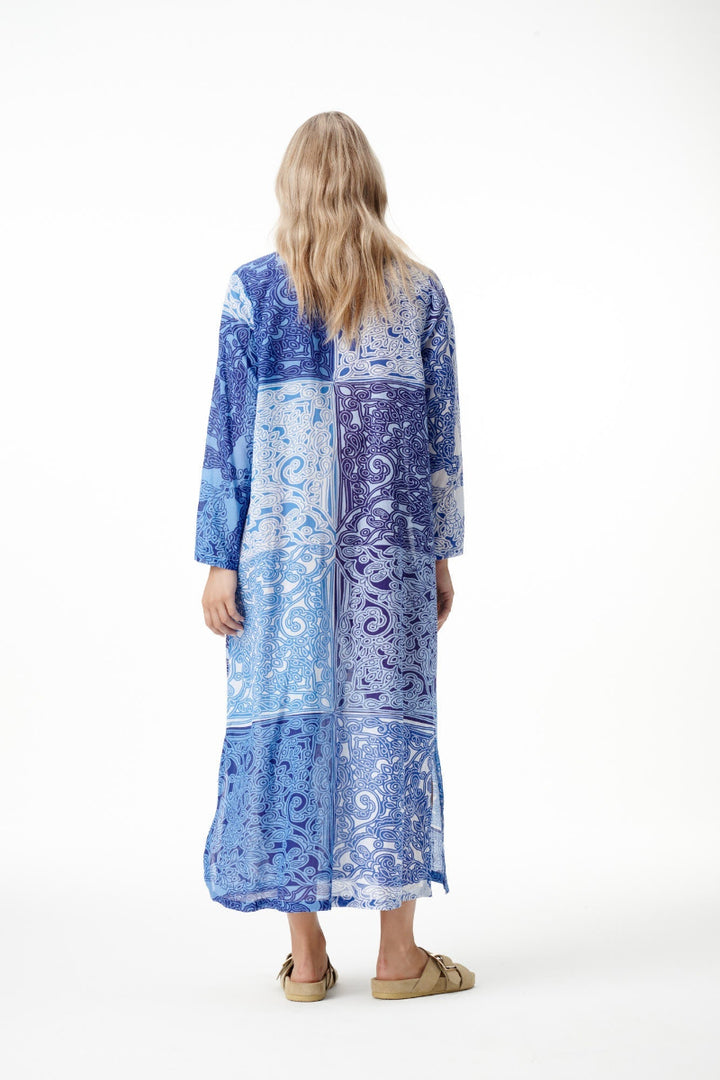 A woman viewed from behind wearing a long blue and white patterned kaftan dress with three-quarter sleeves, paired with tan flat sandals, against a plain white studio background.