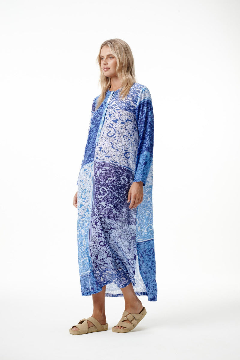 A woman modeling a long-sleeved, loose-fitting kaftan dress with a blue and white paisley pattern, paired with beige slide sandals, against a plain white studio background.