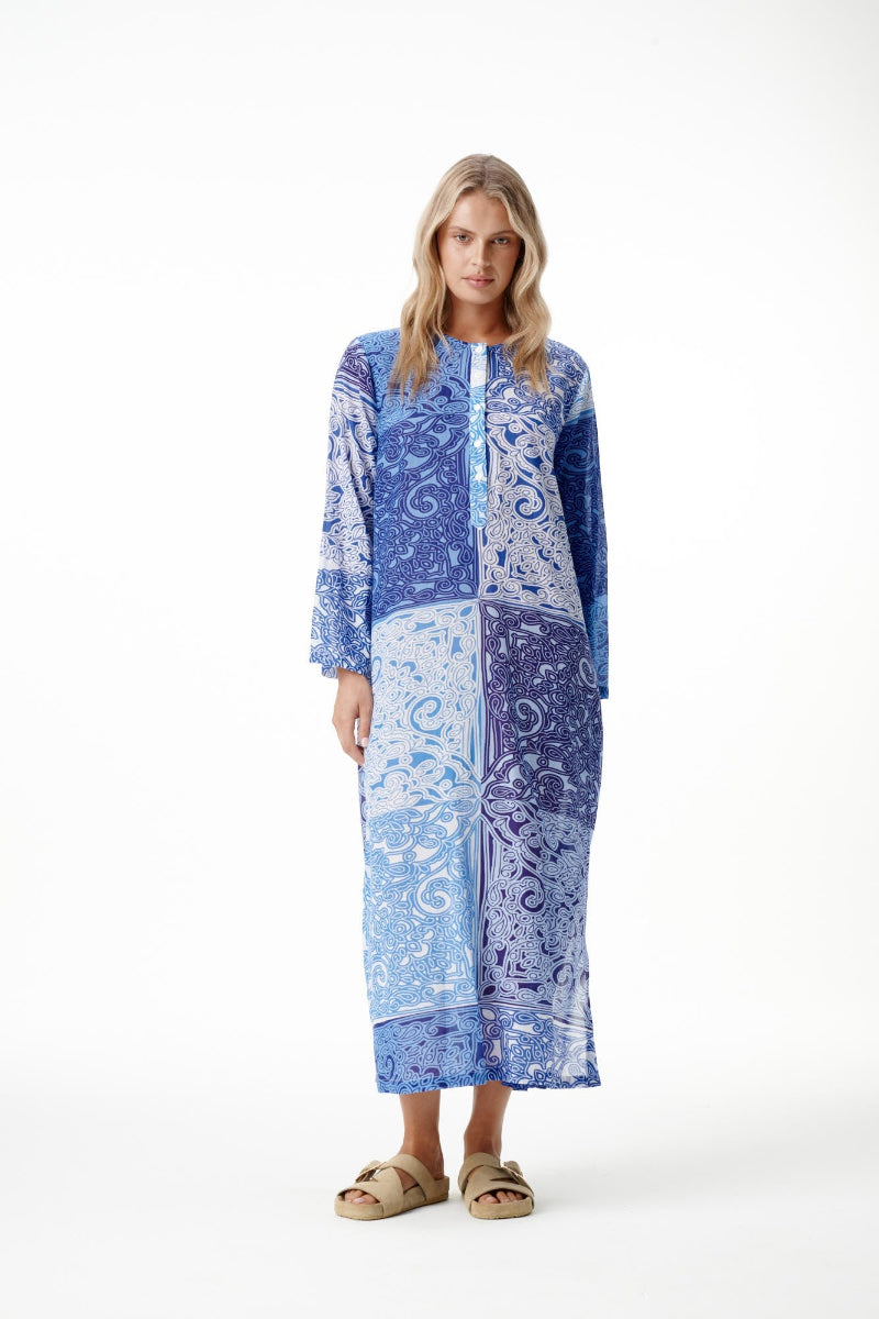 A woman modeling a long-sleeved maxi kaftan dress with a blue and white abstract pattern, standing against a plain white studio background.
