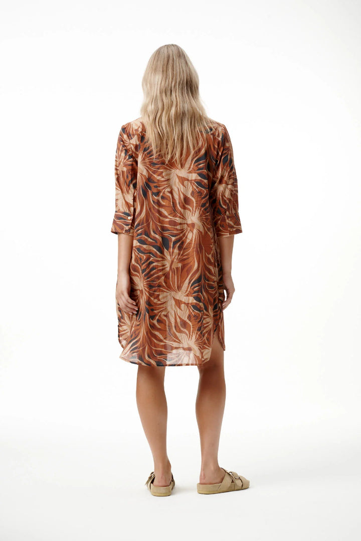 A woman viewed from behind wearing a knee-length brown and beige tropical leaf print tunic dress with three-quarter sleeves, paired with tan slide sandals, against a plain white studio background.