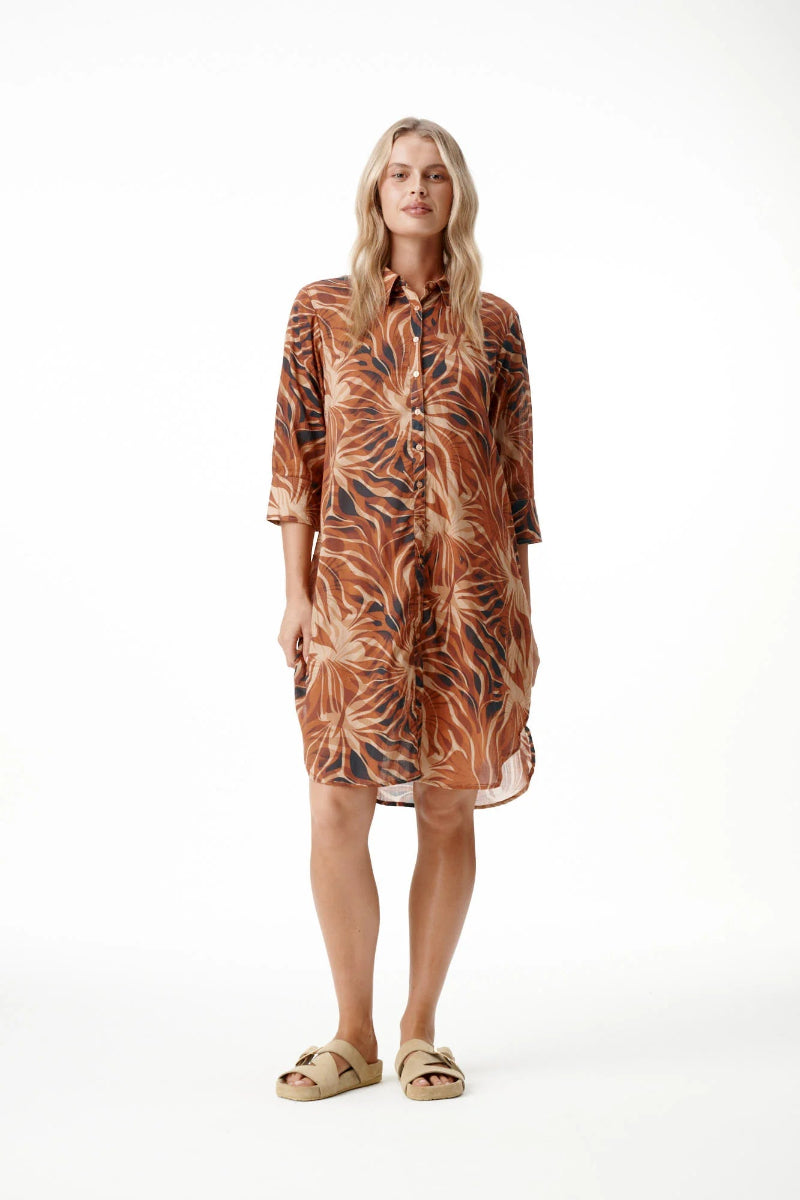A woman modeling a brown and orange tropical leaf print shirt dress with three-quarter sleeves, standing against a plain white studio background.